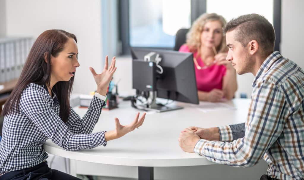 In an office, a woman gestures as she discusses workers’ compensation mediation with a man while another woman looks on.