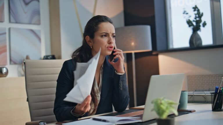Businesswoman talks on the phone about workers’ compensation mediation, holding papers at her desk with a laptop.