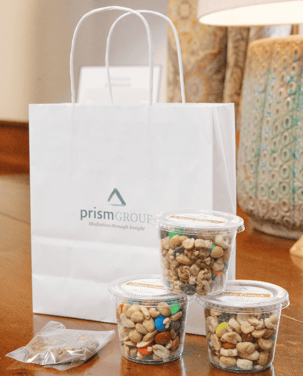 A white Prism Group gift bag sits behind snack cups on a wooden table, ready for a break during mediation services.