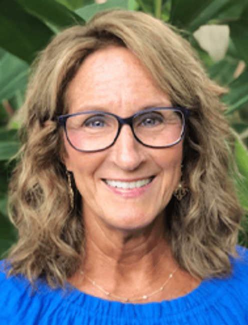Smiling woman with wavy blonde hair, glasses, and a blue top stands before green plants; she is a workers’ comp mediation specialist.