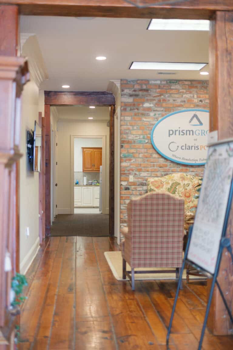 A hallway with wooden floors, a business sign, chairs, and a glimpse into a bright back room used for mediation services.