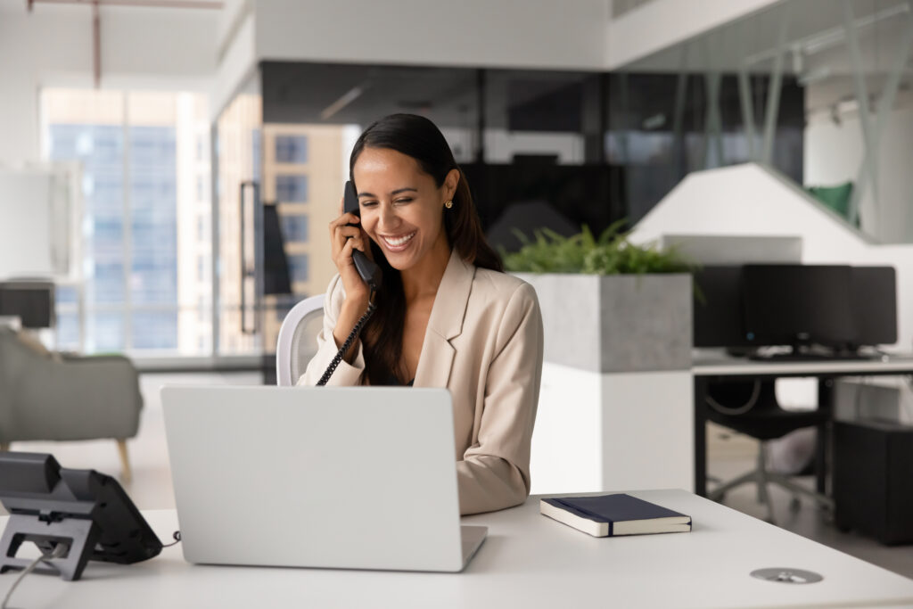 Smiling woman in a beige blazer multitasks at her office desk, on a call about workers’ compensation and working on her laptop.