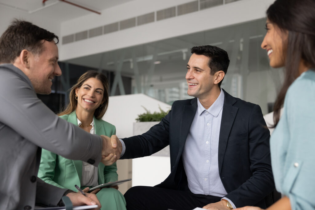 Four professionals in business attire meet for workers’ compensation mediation; two men shake hands as two women smile.