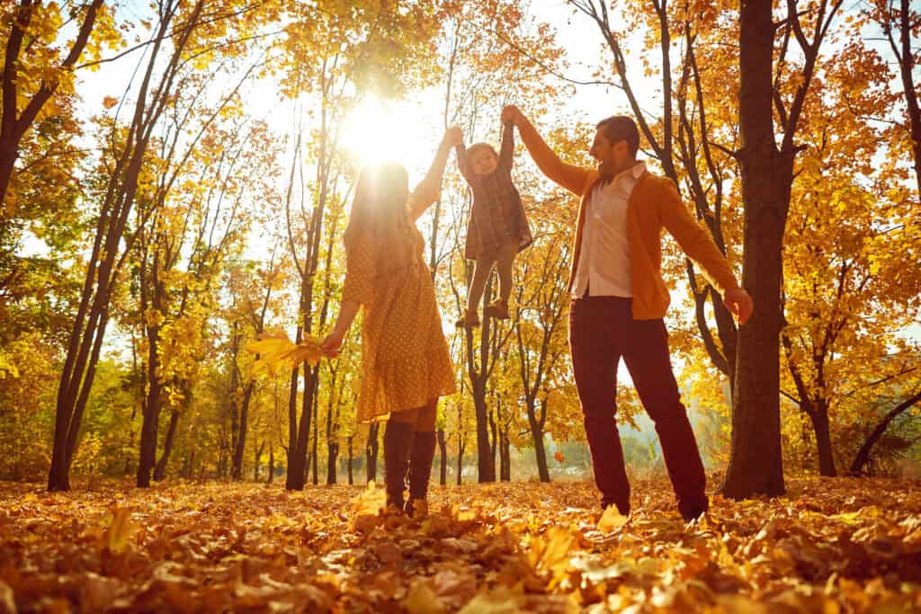 Two adults lift a child in a sunlit forest with autumn leaves, reflecting the supportive nature of legal mediation services.
