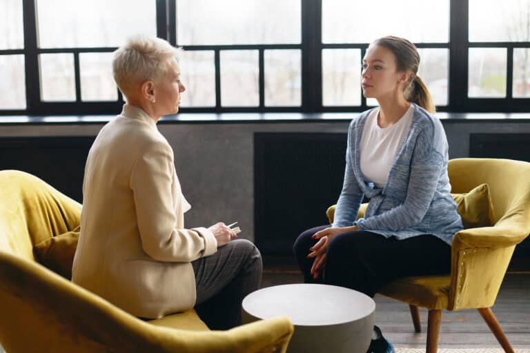 Two women talk about workers’ compensation mediation while sitting in yellow chairs near sunny windows in a bright room.