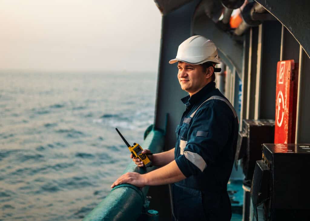 Man in a hard hat and coveralls stands on a ship deck, holding a radio, ready to help with harbor claims mediation.