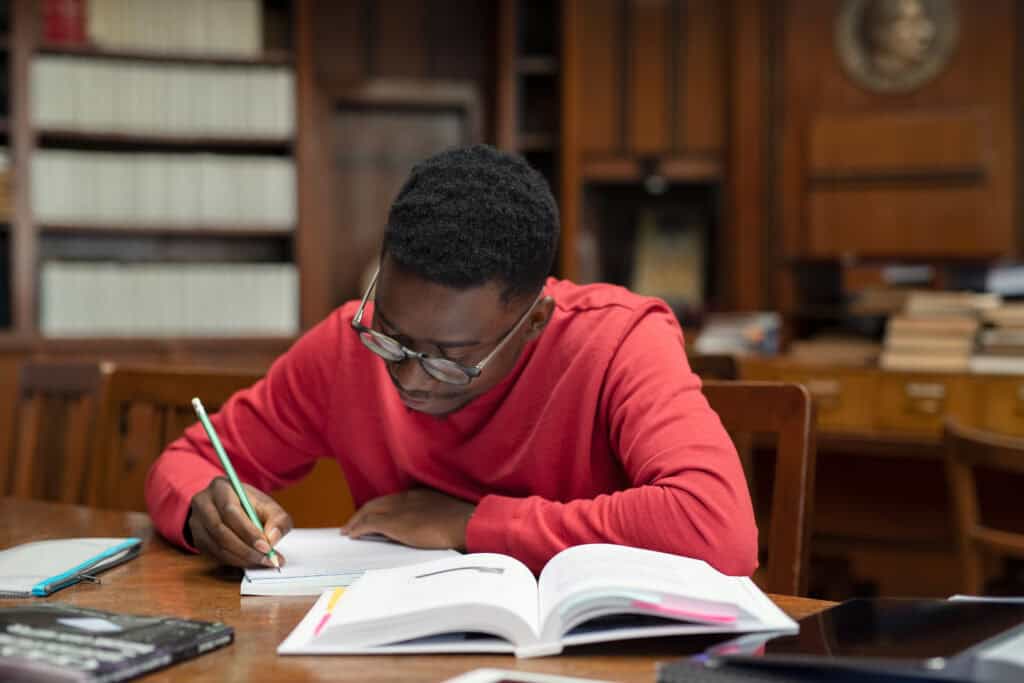 A person in a red shirt writes in a notebook at a library table, researching workers’ compensation mediation.