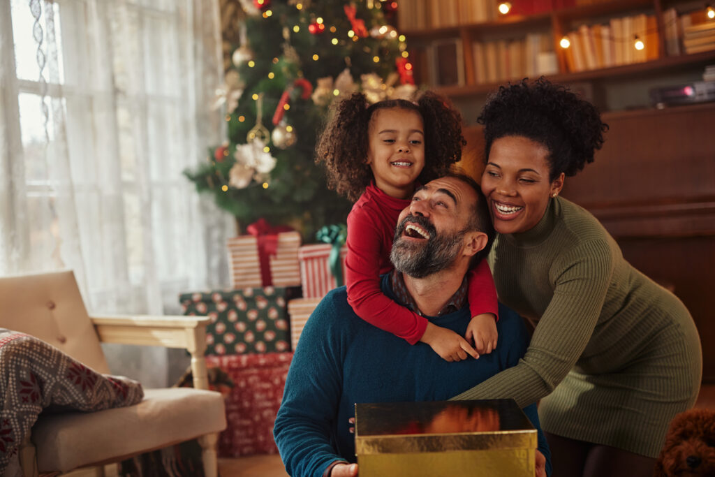A smiling family of three poses by a Christmas tree with gifts. The father, skilled in legal mediation, holds a gold present.