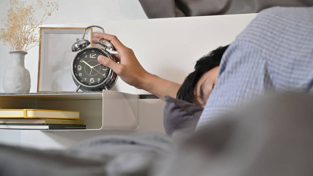 A person in bed reaches to turn off a black alarm clock, with books and a vase nearby—a quiet moment before mediation services begin.