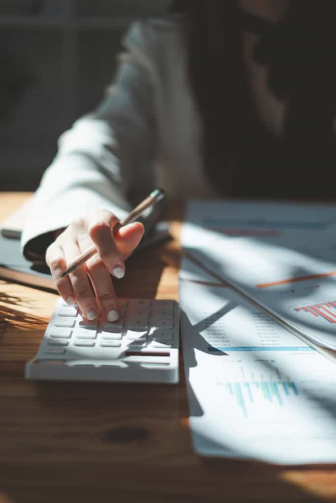 A person calculates with one hand while holding a pen beside workers’ compensation mediation charts on a wooden desk.