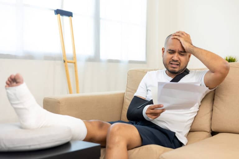 A man with a cast and sling sits on a couch, stressed over paperwork, crutches by his side, seeking workers’ compensation mediation.