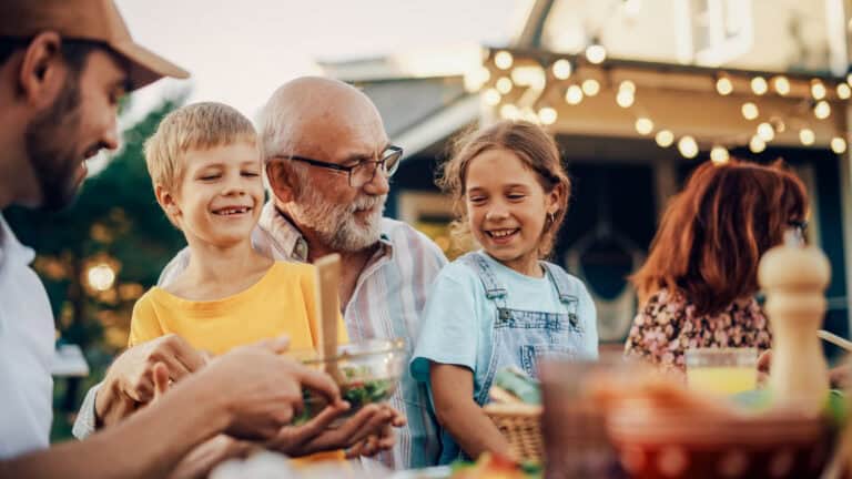 An older man shares a meal outdoors with smiling kids under string lights, enjoying a calm break from workers’ comp mediation.