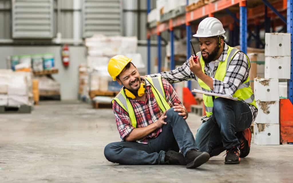 A worker clutching his knee on a warehouse floor in pain while a colleague calls for help, illustrating workers’ comp mediation.