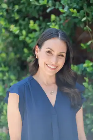Smiling woman with long brown hair in a blue blouse stands outdoors, prepared to help with workers’ compensation mediation services.