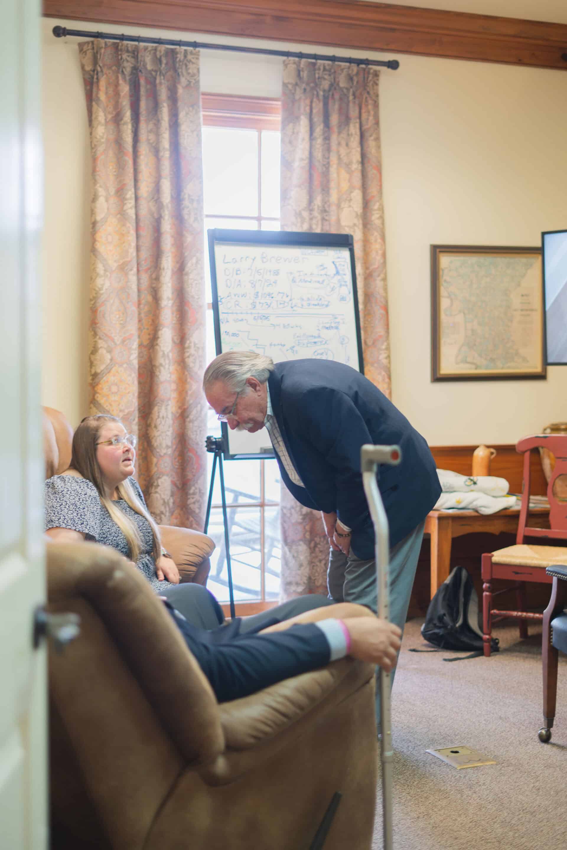 A man leans toward a seated woman in an office, possibly discussing workers’ compensation mediation near a whiteboard and cane.