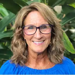 Smiling woman with wavy light brown hair, glasses, and a blue top stands before green plants; she mediates workers’ compensation cases.