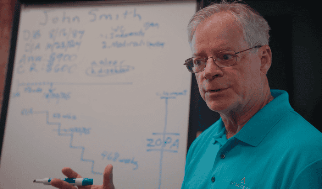An older man in glasses and a teal shirt explains workers’ compensation mediation by a chart-filled whiteboard.