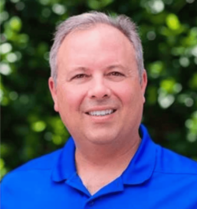 A smiling man with short gray hair stands before blurred green foliage, symbolizing workers’ compensation mediation.