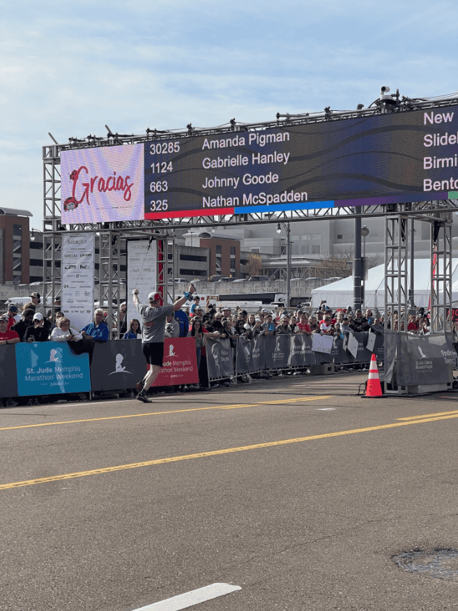 A runner celebrates at the marathon finish line, with spectators and a digital board showing race dispute mediation services.