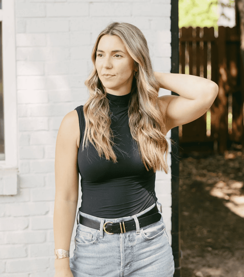 A woman with long, wavy hair in jeans stands by a white brick wall, contemplating workers’ compensation mediation.