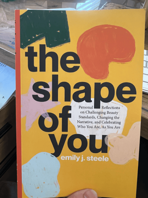 A hand holds “The Shape of You” by Emily J. Steele, featuring a bold yellow cover with abstract shapes—great for mediation inspiration.
