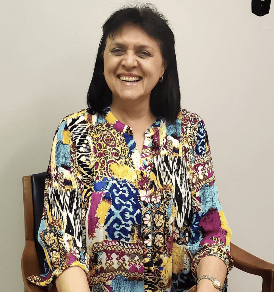 A woman with dark hair in a bright blouse smiles warmly, conveying confidence in legal mediation, standing by a light wall.