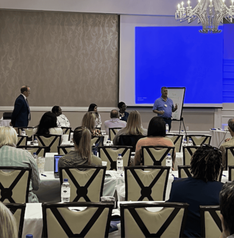 A man presents on workers’ compensation mediation at a flip chart, with attendees and a blue projector screen nearby.