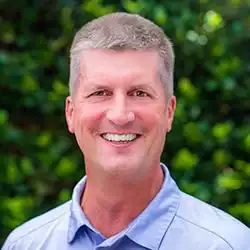Smiling man with short gray hair in a light blue shirt stands before green foliage, ready to help with mediation services.