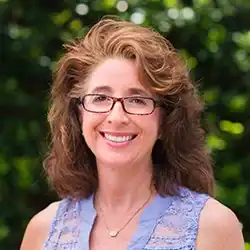 Smiling woman with wavy brown hair and glasses, in a light purple blouse, legal mediation expert amid green foliage.