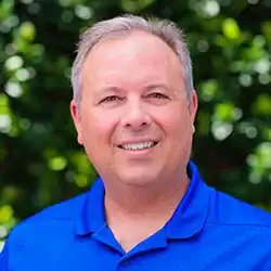 A smiling man with short gray hair and a blue polo stands before lush greenery; he specializes in workers’ compensation mediation.