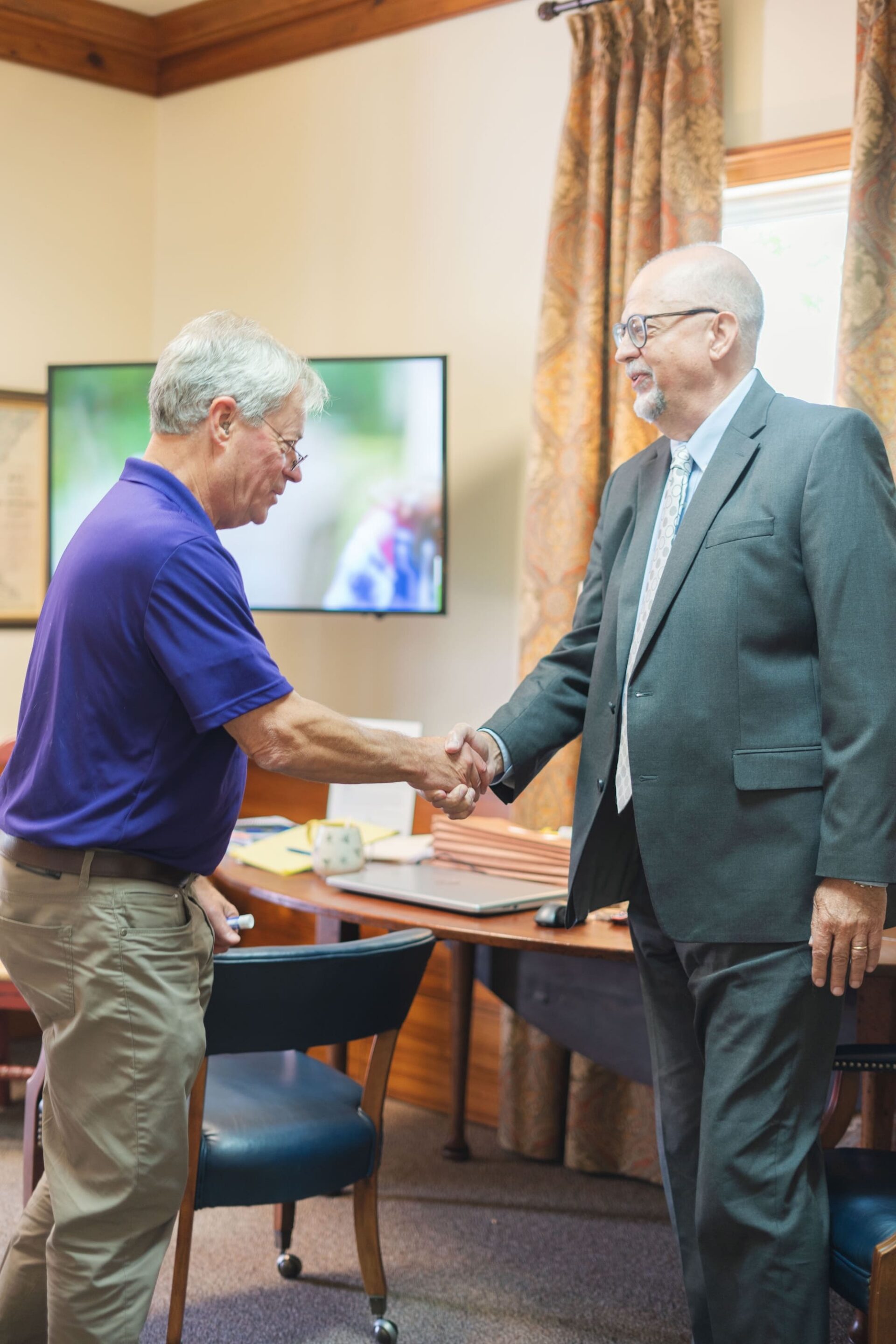 Two men, one in a suit and one in a polo, shake hands in an office as workers’ compensation mediation experts.