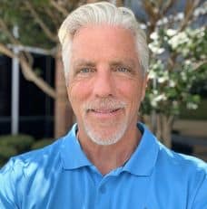 Smiling older man with white hair and a goatee, wearing a light blue polo outdoors; skilled in legal mediation.