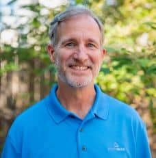 Smiling gray-haired man with a beard in a blue polo stands outdoors before trees, skilled in workers’ compensation mediation.