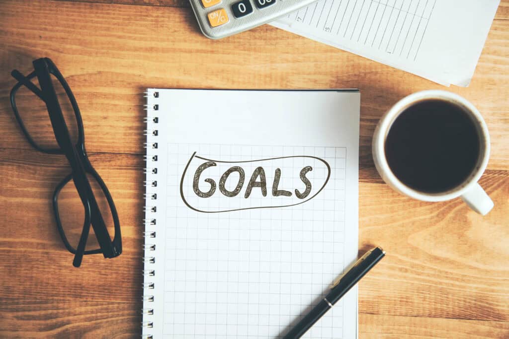 A notebook labeled “GOALS,” pen, eyeglasses, coffee, and a calculator sit together on a wooden desk for mediation work.