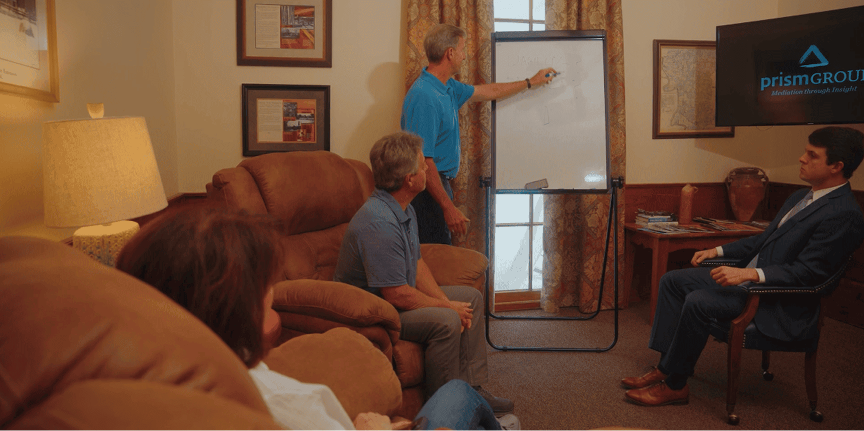 A man in a blue shirt discusses workers’ compensation mediation with three people in a cozy office, TV shows prismGROUP logo.