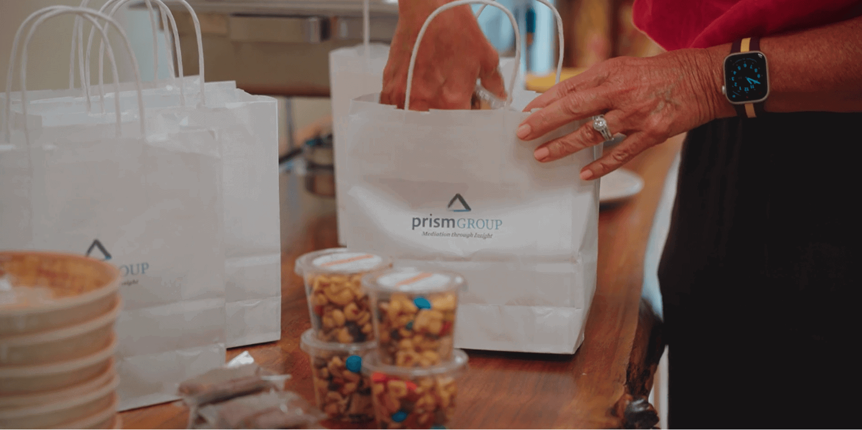 A person with a smartwatch packs items into a Prism Group gift bag at a table with snacks, promoting mediation services.