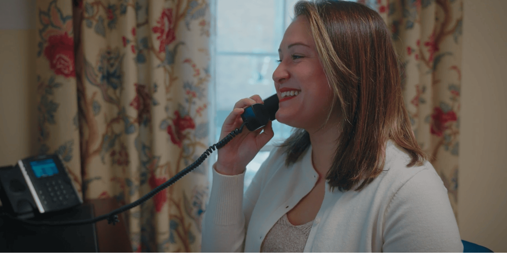 A woman in a white sweater smiles while talking on a corded phone about workers’ comp mediation, framed by floral curtains.