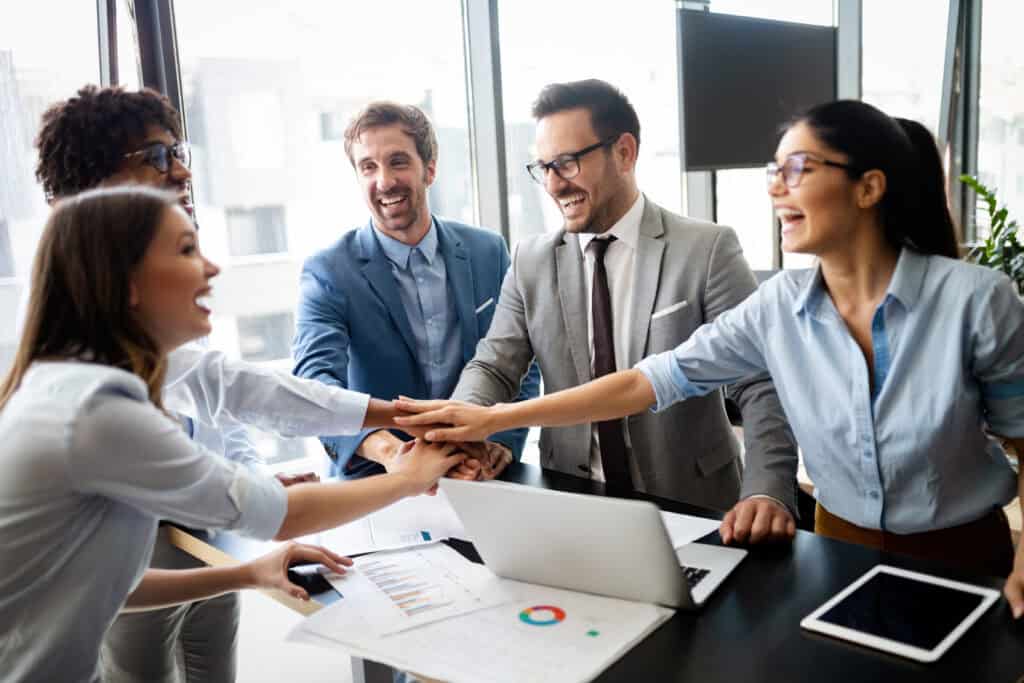 Five colleagues stack hands over laptops and papers, showing teamwork during a workers’ compensation legal mediation in a modern office.