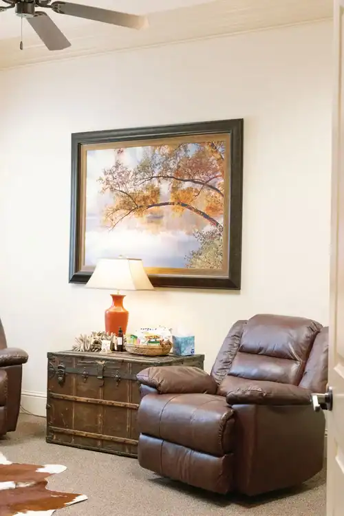 A brown leather recliner beside a lamp and chest sits under a tree painting, perfect for quiet legal mediation discussions.