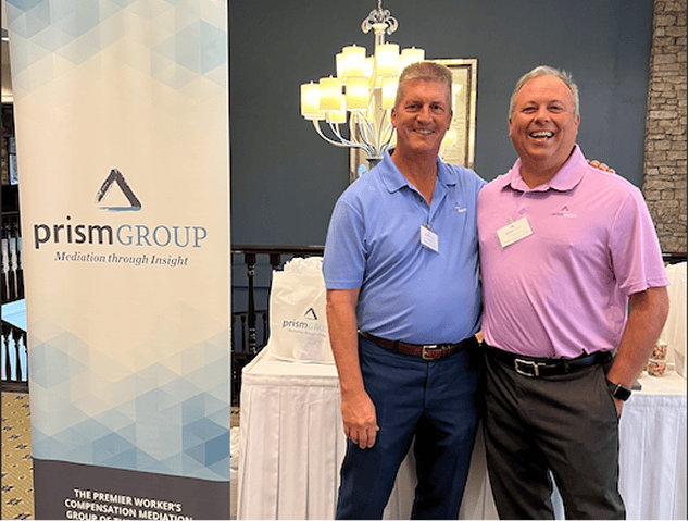 Two men smile next to a Prism Group banner at an event focused on workers’ compensation legal mediation services.