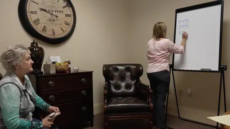 Two women talk about workers’ compensation mediation by a whiteboard labeled “TTD, PTD, PPD” in an office meeting.