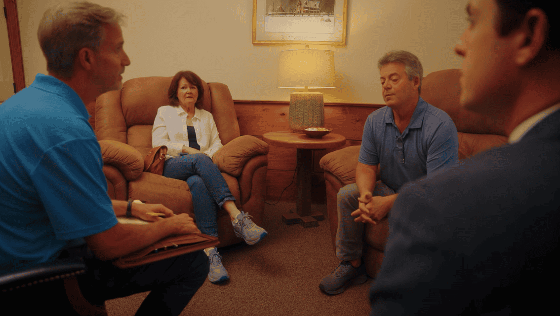 Four adults in a warmly lit room participate in mediation; one, holding a clipboard, appears to guide the discussion.