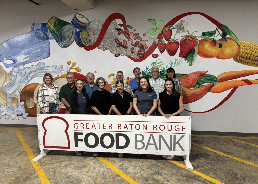 Thirteen people pose behind a “Greater Baton Rouge Food Bank” sign in front of a mural, supporting legal mediation services.