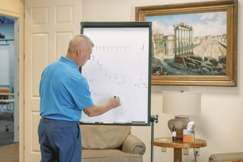 A man in a blue shirt explains workers’ compensation mediation, writing on a whiteboard in an office.
