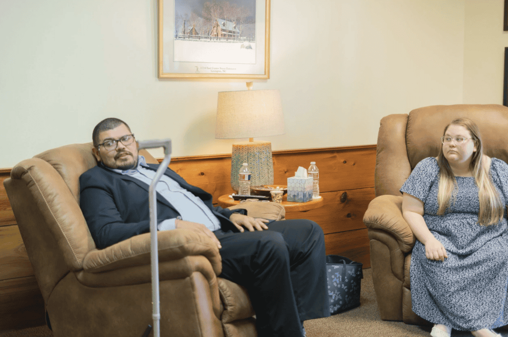 A suited man and a woman in blue talk about workers’ compensation mediation in a warmly lit wood-paneled room with art.