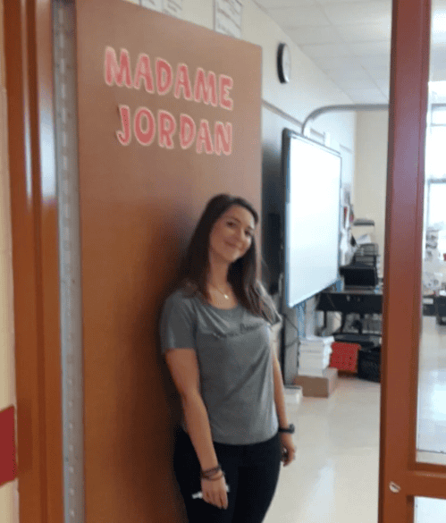 A woman stands by a classroom door labeled “Madame Jordan,” with desks and technology inside for legal mediation discussions.
