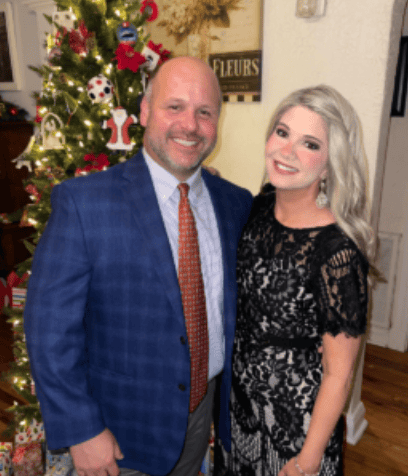 A man in a blue suit and a woman in a black lace dress, both mediation professionals, smile by a decorated Christmas tree.