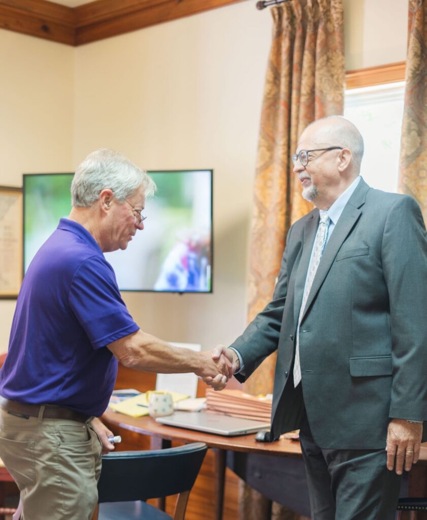 Two men, one in a suit and one in a polo, shake hands in an office as workers’ compensation mediation experts.