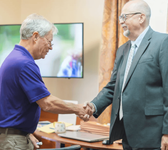 Two men shake hands in an office after a legal mediation; a desk and TV can be seen in the background.