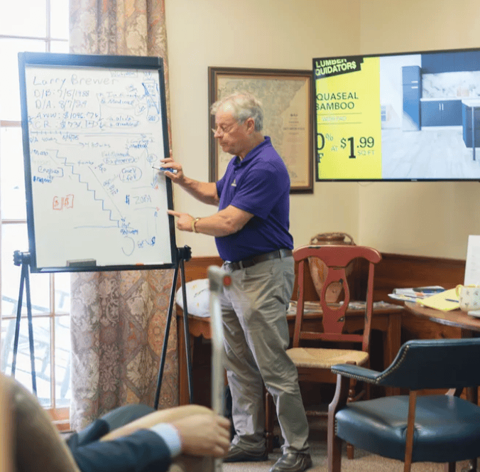 An older man in a purple shirt writes on a whiteboard while a legal mediation ad is displayed on the TV behind him.